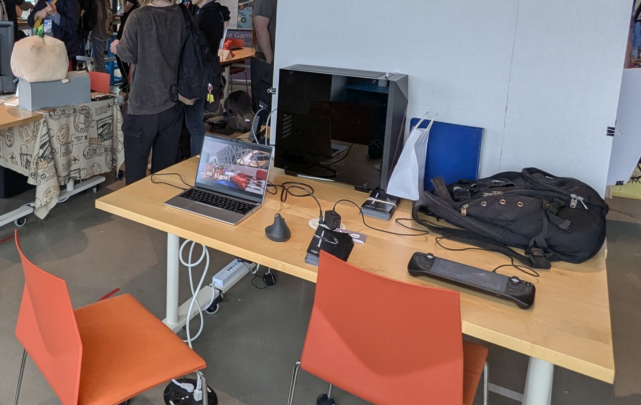 My desk at the GodotCon Boston showcase, which is a bare table with flyers, business cards, a laptop with a mouse, and a steam deck on it. My backpack sits behind my stuff. There's two orange seats in front of the table.